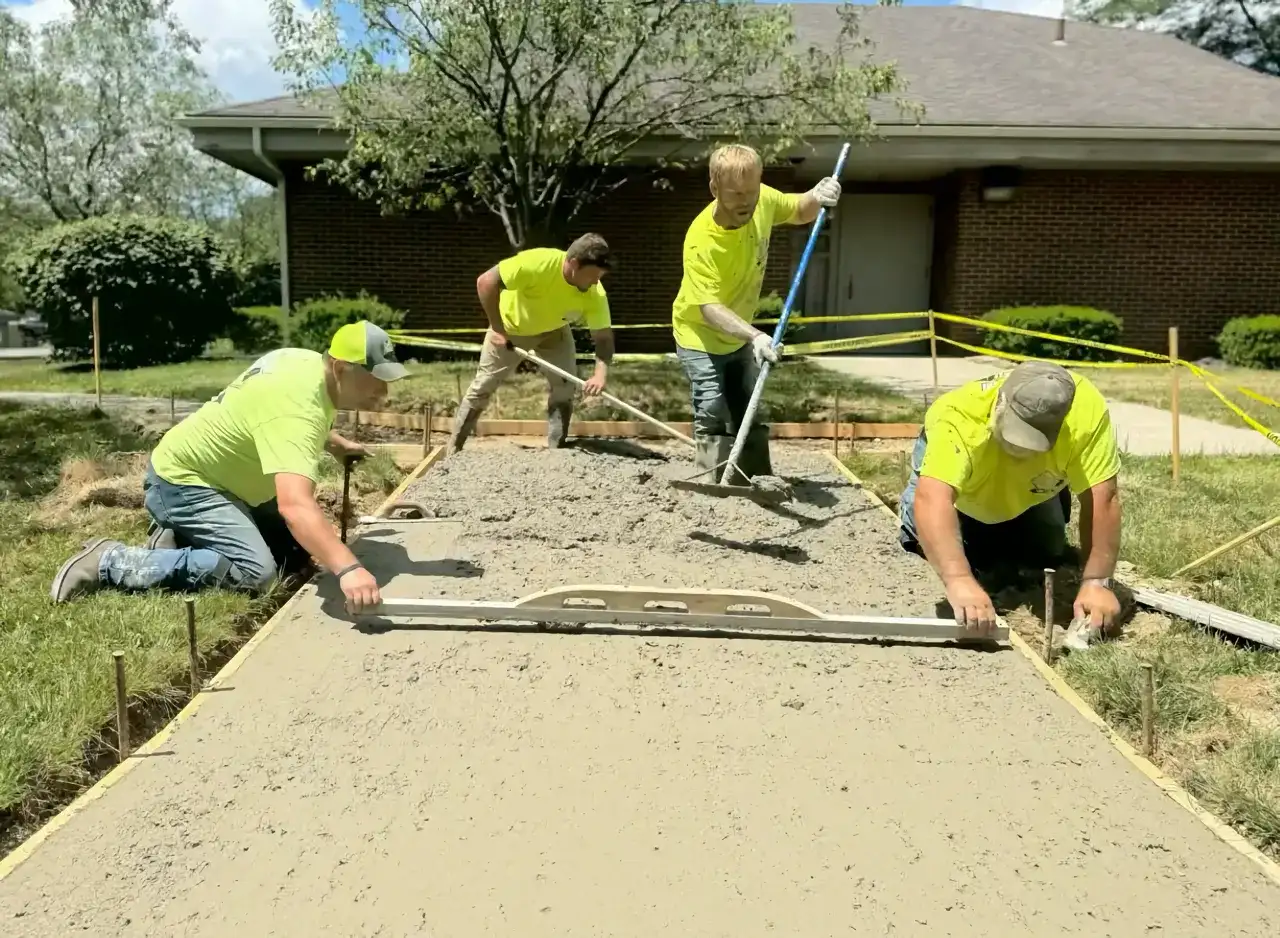 Four men in yellow shirts screeding a fresh concrete walkway between staked forms beside a brick building