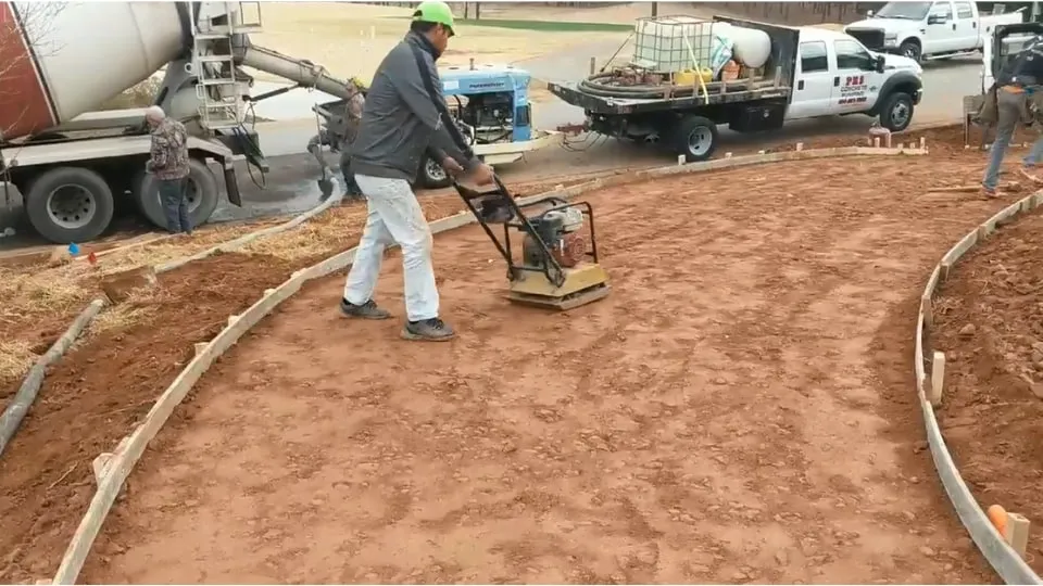 A worker running a plate compactor over red clay inside curved driveway forms, with a mixer truck behind